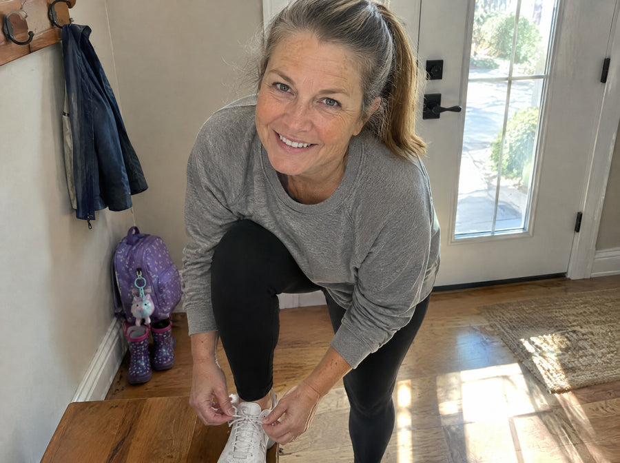 A woman around 56 bending easily to tie her shoes in a sunny hallway, smiling